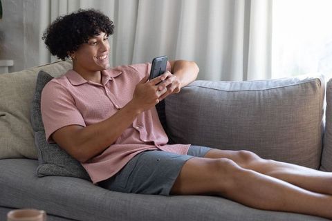 Smiling young man reclining on gray sofa using smartphone for messaging and streaming