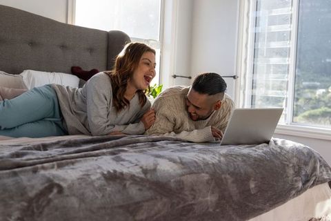 Couple laughing and streaming on laptop while relaxing on bed in sunlit cozy bedroom