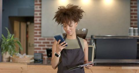African american barista checking smartphone holding tablet at rustic coffee counter