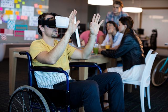 Man in wheelchair using VR headset in modern office with colleagues in background. Ideal for illustrating themes of accessibility, inclusion, technology in the workplace, and innovative work environments.