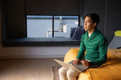Asian man working remotely on laptop from bed edge beside window bench, mustard blanket