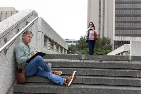 Middle-aged man using tablet on urban concrete steps with coffee while commuter walking by