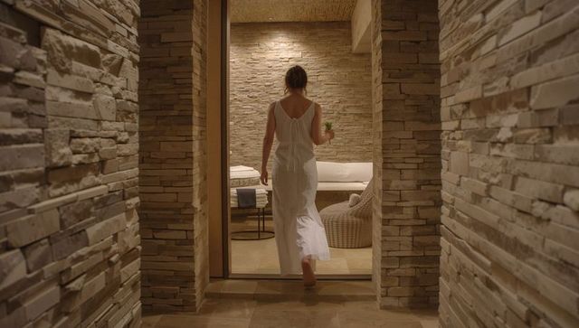 Barefoot woman walking into minimalist stone spa lounge carrying potted plant wellness