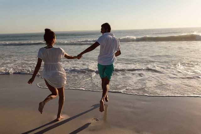 This image depicts a couple holding hands and running along the shoreline at sunset. The scene exudes romance and joy, making it perfect for use in travel advertisements, romance-themed content, summer vacation promotions, and lifestyle blogs. The warm, golden light enhances the romantic and carefree atmosphere.