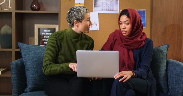 Two young women, one Caucasian with a green turtleneck and the other Middle Eastern wearing a hijab, are focused on a laptop screen, with copy space. They could be colleagues or students collaborating on a project or discussing work.