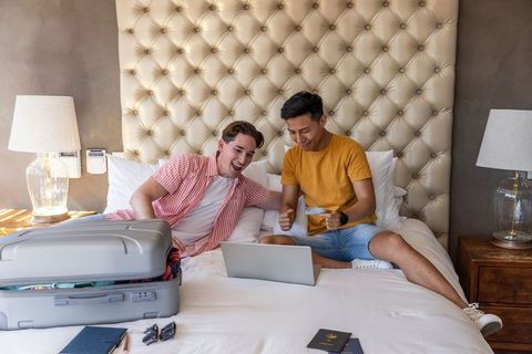 Diverse male friends packing suitcase and booking flights on laptop in hotel room