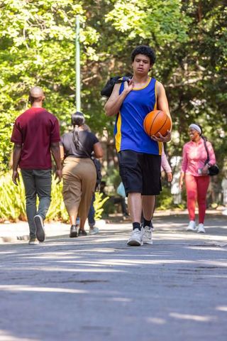Young man carrying basketball walking through shaded campus park with sports bag