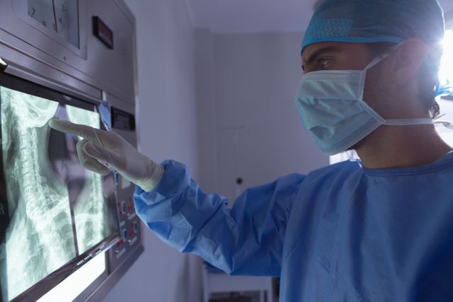 This image shows a male surgeon in an operation room, closely examining an x-ray on a light box. He is wearing protective gear including a surgical mask, cap, and gloves. This image can be used in medical articles, healthcare websites, educational materials, and hospital brochures to illustrate medical examinations, surgical procedures, and healthcare professionals at work.