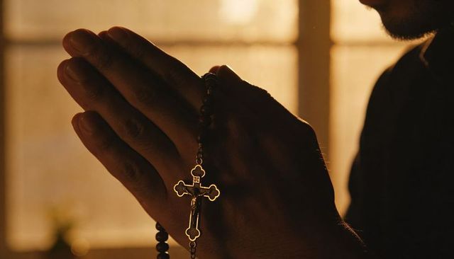 Praying hands holding rosary and crucifix backlit by warm window light