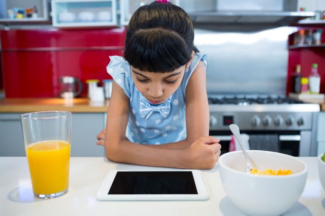 Young girl focused on tablet while having breakfast in a contemporary kitchen. Ideal for use in educational content, technology in daily life, family lifestyle, and modern home settings.