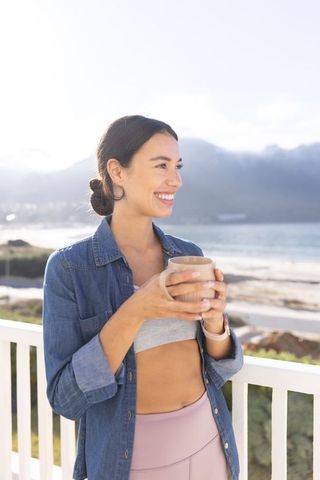 Smiling woman holding coffee mug on balcony overlooking beach sunrise wellness lifestyle