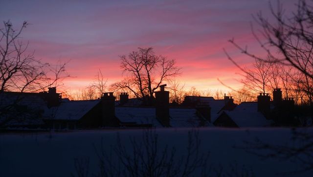 Winter dawn silhouette of snow-covered rooftops and chimneys against pink purple sky glow