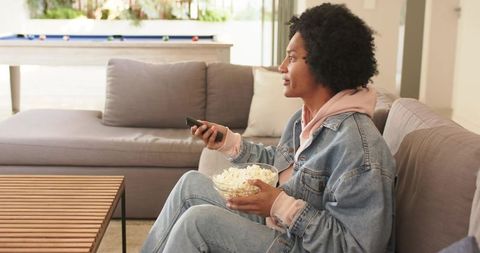 30s african american man relaxing on couch watching tv holding popcorn bowl and remote