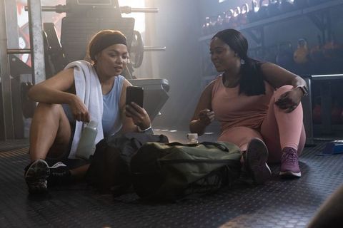 Two women resting after workout sharing phone and laughing in gym with kettlebells