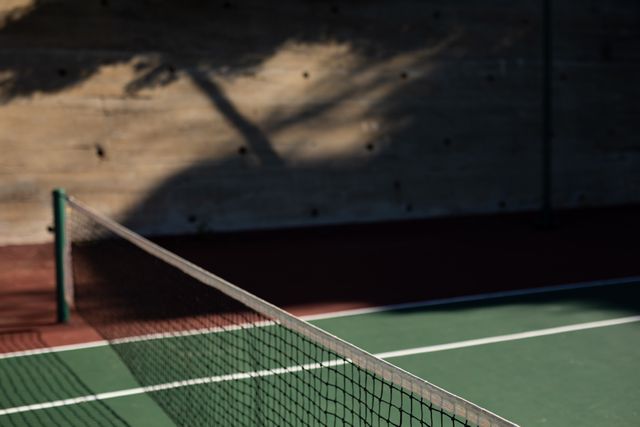 General view of an empty tennis court with a net in the middle on a sunny day with a shadow of a tree on a wall in the background.
