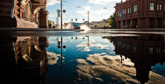 Urban scene captures car driving by buildings reflected in puddle on clear day, showcasing city architecture and movement. Ideal for urban lifestyle themes, city planning, weather effects, transportation, and reflections in photography.