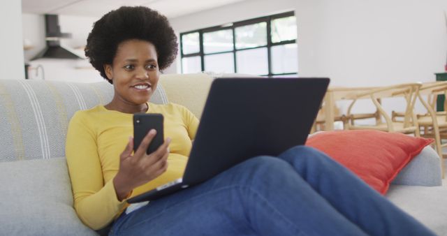 African American woman sitting comfortably on a couch with a laptop on her lap and holding a smartphone, smiling and looking relaxed. Ideal for depicting remote work, technology use, daily life at home, and casual relaxation scenes. Useful for advertisements, blog posts, articles on modern technology use in everyday life, and social media content showing a balanced lifestyle.
