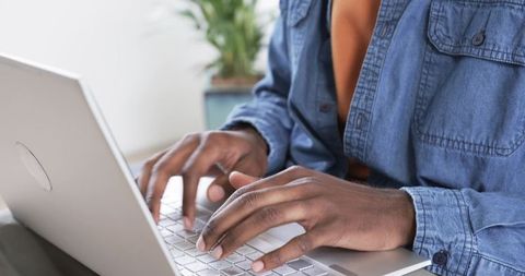 African American man typing on laptop at home wearing denim shirt and orange top close-up