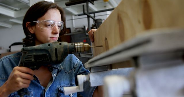 Young Caucasian woman drills into wood at a workshop. Safety goggles on, she focuses intently on her carpentry project.