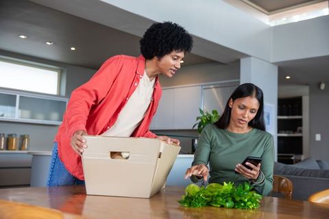 Couple Unpacking Box with Fresh Greens and Smartphone in Kitchen