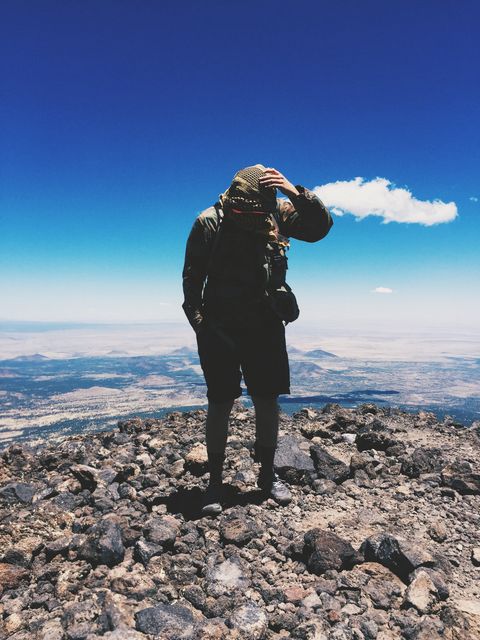 Hiker shielding face from sun standing on mountain peak wearing outdoor gear. Blue sky backdrop with expansive landscape view. Suitable for adventure, hiking, and travel promotions or inspirational content. Perfect for nature blogs, outdoor gear advertisements, or motivational posters.