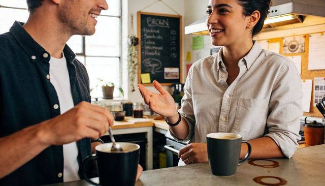 Smiling couple chatting over coffee at cozy sunlit cafe counter, casual button-up style