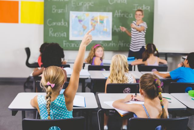 Schoolgirl raising hand in classroom during geography lesson with teacher pointing at map on chalkboard. Students are engaged and participating in the lesson. Ideal for educational content, school promotions, learning materials, and articles on classroom activities.