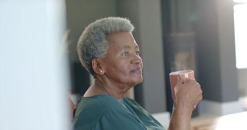 Senior african american woman sipping steaming mug in sunlit living room, serene portrait