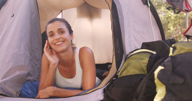 Woman is lying inside a tent, resting and smiling. The scene depicts outdoor camping and the tranquility of nature. There are hiking backpacks visible in the foreground, indicating preparation for hiking or traveling. This content can be used for advertising travel gear, promoting outdoor activities, or illustrating articles on travel and adventure.