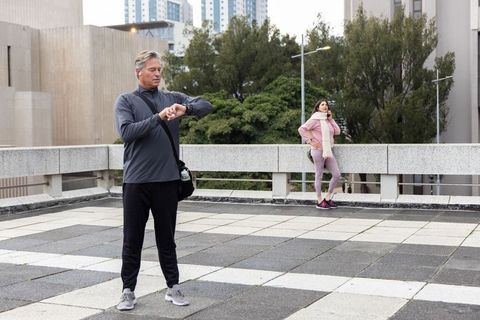 Mature man checking smartwatch on urban rooftop while woman talking on phone, promoting fitness