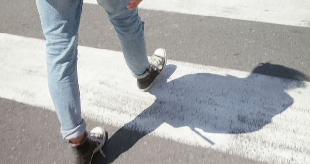 Person crossing a street on a pedestrian crosswalk wearing casual jeans and sneakers. Shadow is visible on the road. Ideal for concepts related to urban lifestyle, pedestrian safety, and transportation.