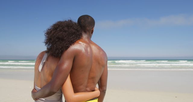 An African American couple enjoys a romantic moment on a sunny beach, with copy space. Embracing each other, they gaze out at the ocean, symbolizing a peaceful and loving relationship.