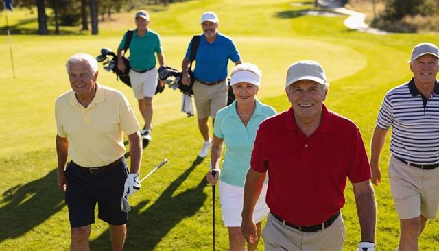 Active senior golfers walking together on sunny course wearing polos, visors, carrying bags