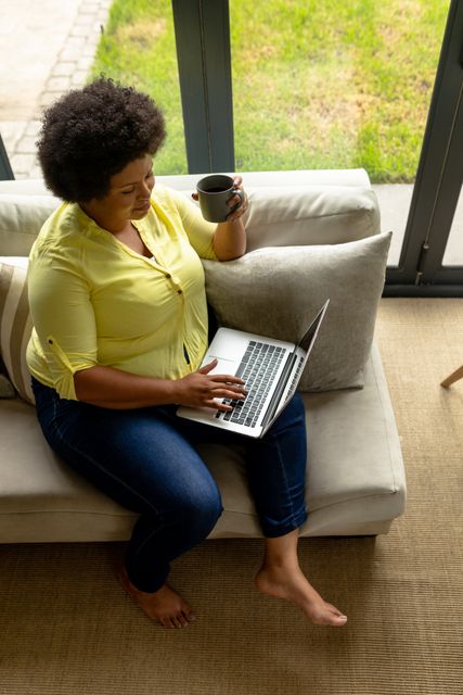 This image shows an African American mid adult woman sitting comfortably on a sofa, using a laptop and holding a cup of coffee. Ideal for illustrating concepts related to remote work, home office setups, relaxation, and modern lifestyle. Suitable for articles, blogs, and advertisements focusing on work-life balance, technology use at home, and leisure activities.