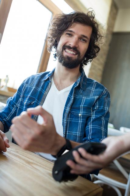 Man smiling while using smart watch for contactless payment at table. Ideal for illustrating modern technology, digital transactions, and lifestyle themes. Suitable for articles on fintech, smart devices, and casual dining experiences.