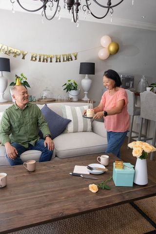 Chinese senior couple celebrating cozy home birthday woman carrying cake man wearing party hat