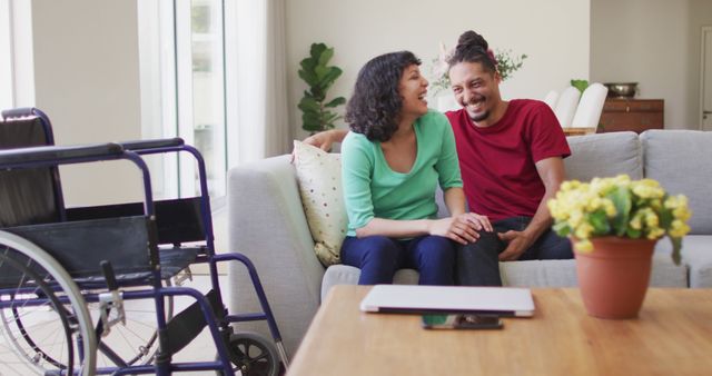 Happy biracial couple sitting on couch laughing in living room, with wheelchair in foreground. wellbeing and domestic lifestyle with physical disability.