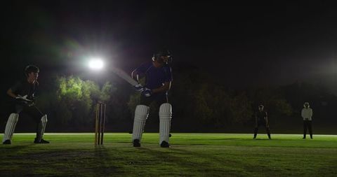 Night cricket batsman batting under floodlights at crease wearing helmet and pads, stumps