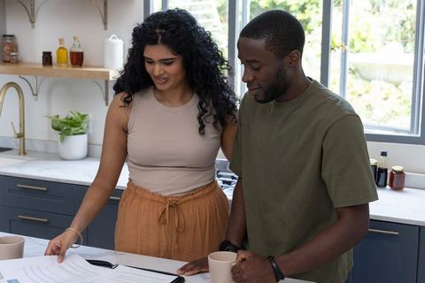Diverse couple standing in modern kitchen reviewing documents and sharing coffee