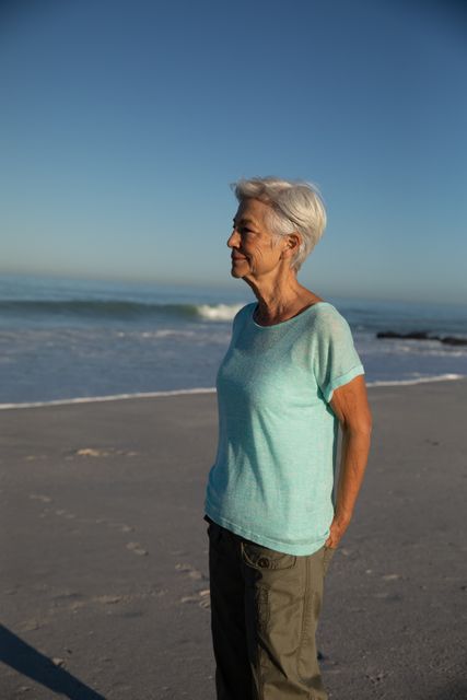 Senior Caucasian woman enjoying time at the beach on a sunny day, standing on the beach, with sea and blue sky in the background. Summer tropical beach vacation.