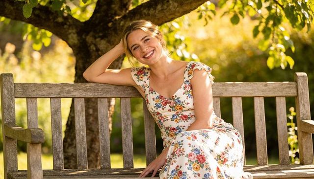 Smiling woman relaxing on wooden bench in sunlit garden wearing vintage floral summer dress