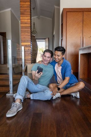 Multiracial partners sitting on hardwood floor sharing smartphone in modern home