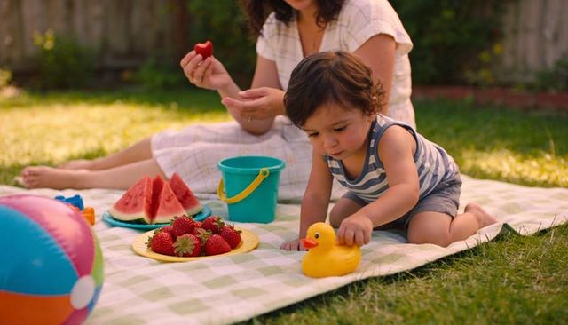 Curious toddler reaching for yellow rubber duck during backyard picnic with strawberries on blanket