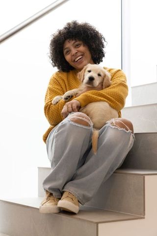 Smiling woman sitting on stairs holding golden retriever puppy, cozy home casual lifestyle