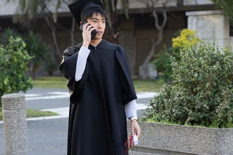 Asian graduate wearing mortarboard and gown talking on smartphone while holding diploma