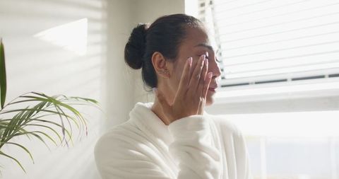 Mid-adult asian woman applying skincare by sunlit bathroom window in white plush robe