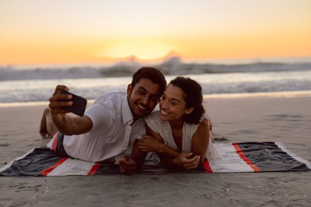 Couple lying on beach blanket taking selfie with mobile phone at sunset. Ideal for use in travel promotions, romantic getaway advertisements, social media content, and lifestyle blogs showcasing love and relaxation.