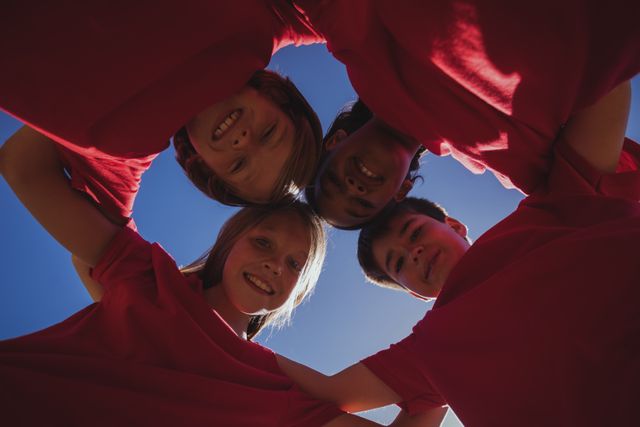 This image shows a group of happy kids forming a huddle during a boot camp. They are wearing red shirts and smiling, symbolizing teamwork, unity, and friendship. This image can be used for promoting summer camps, team-building activities, educational programs, and youth events.