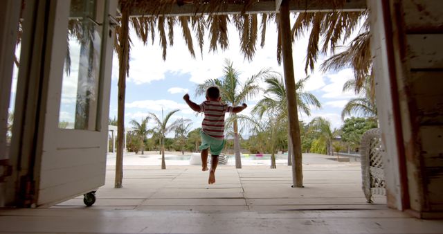 Happy caucasian boy running and raising hands at swimming pool at beach house with palm trees. Vacation, free time, summer and childhood.