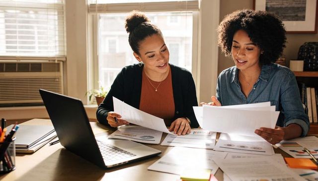 Two women reviewing charts and laptop, collaborating on business planning in sunlit office